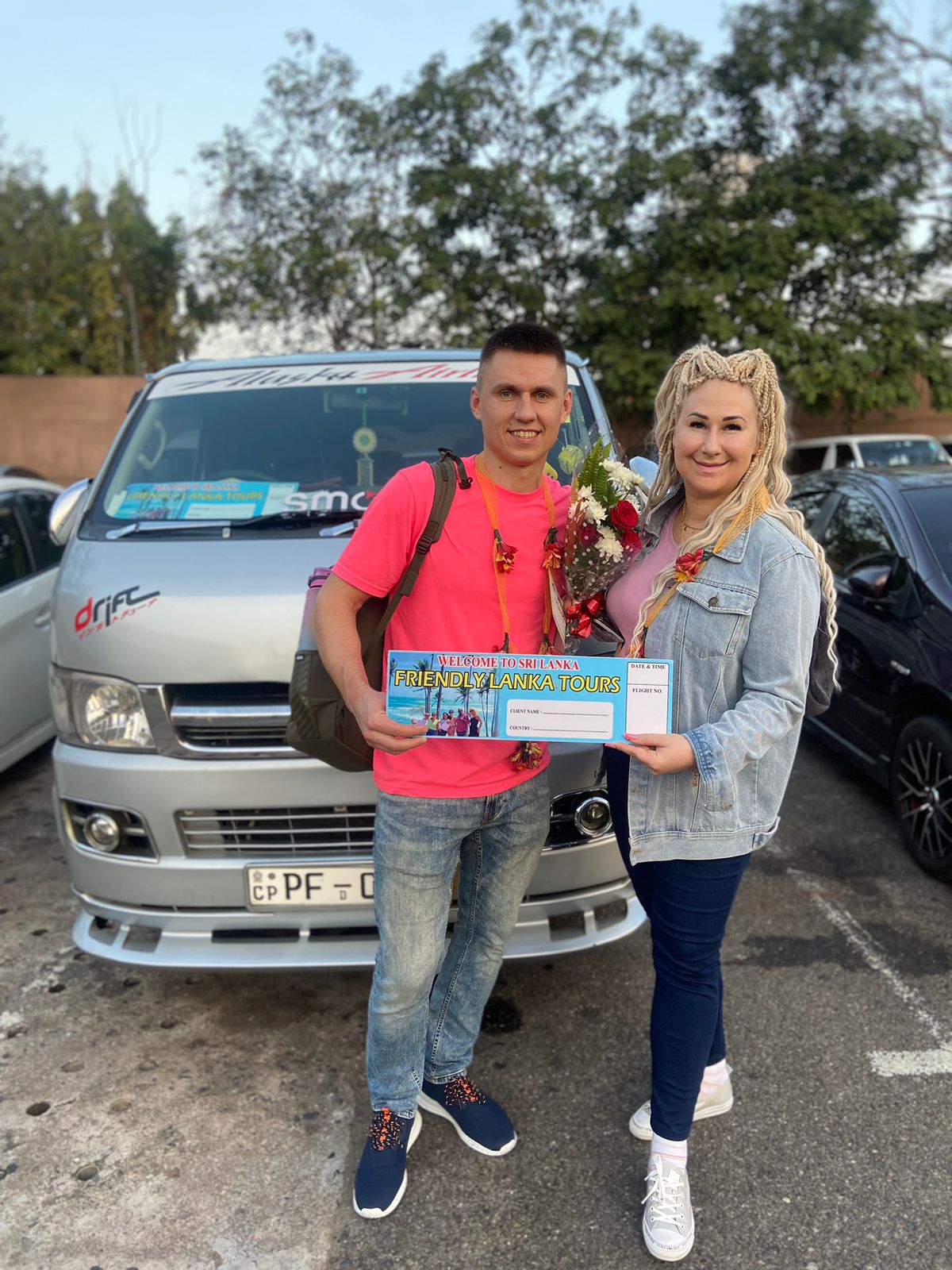 Two travellers standing with a welcome sign beside a van